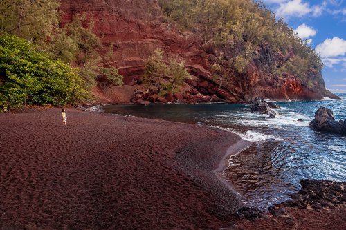 A person stands on a red sand beach, surrounded by lush greenery and rocky cliffs, with waves gently lapping at the shore.