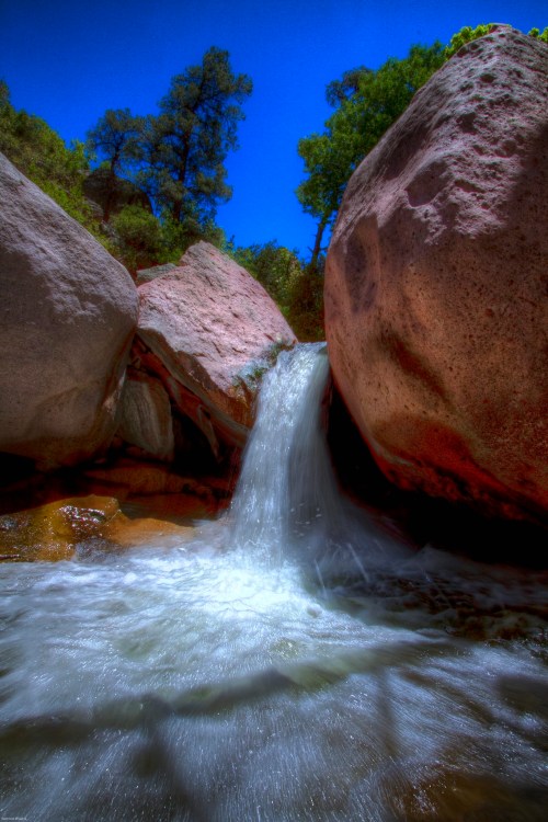 A small waterfall cascades between large rocks, surrounded by lush greenery and a clear blue sky.