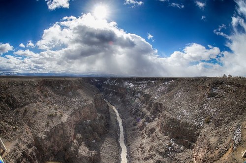 A panoramic view of a deep canyon with a river, under a bright sun and partly cloudy sky. Snow is visible on the ground.