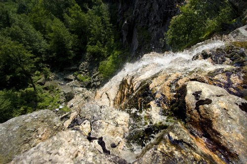 A cascading waterfall flows over rocky terrain, surrounded by lush green trees and a steep cliff.