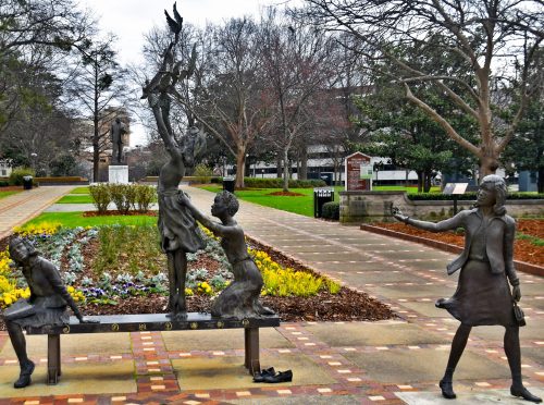 Three bronze sculptures of children playing in a park, surrounded by colorful flowers and trees.
