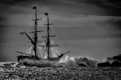 A silhouette of a tall ship sailing through rough waters, with waves crashing against its hull under a moody sky.
