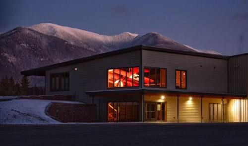 A modern building with large windows, illuminated from within, set against a snowy mountain backdrop at dusk.
