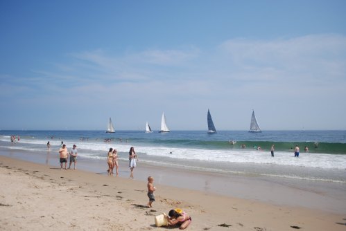 A sunny beach scene with people swimming, playing, and several sailboats in the distance on calm waters.