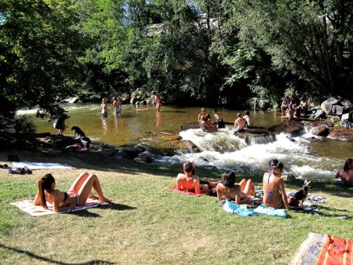 People enjoying a sunny day by a river, some swimming, others sunbathing on the grass. Trees and rocks in the background.