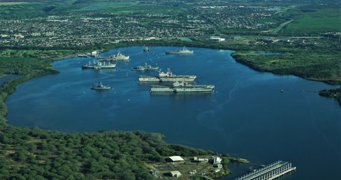 Aerial view of several naval ships anchored in a calm bay, surrounded by greenery and a distant urban area.