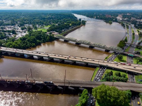 Aerial view of a river with bridges, highways, and lush greenery along the banks under a cloudy sky.