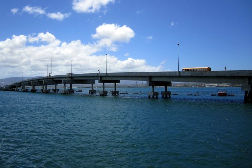 A long bridge stretches over blue water, with a bus on the roadway and clouds in a clear sky above.