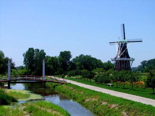 A scenic view of a windmill beside a canal, with a small bridge and lush greenery in the background.