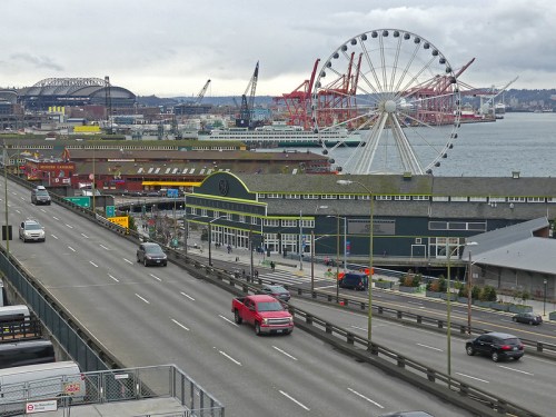 A view of a waterfront with a Ferris wheel, shipping cranes, and cars on a road under a cloudy sky.