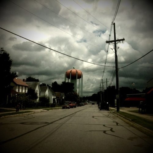 A street view with a water tower resembling a giant orange, under a cloudy sky, lined with power poles and houses.