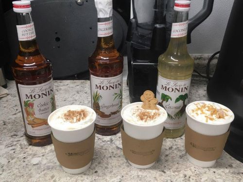 Three cups of festive drinks topped with whipped cream and garnishes, alongside bottles of Monin syrup: Cookie Butter, Gingerbread, and Coconut.
