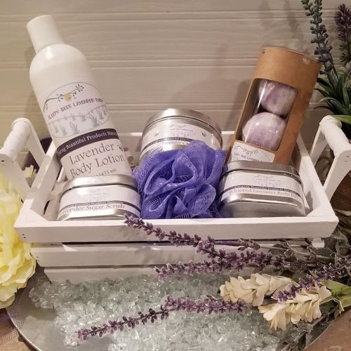 A white basket filled with lavender body lotion, scrubs, bath bombs, and a loofah, surrounded by lavender flowers.