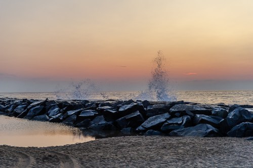 Waves crashing against a rocky jetty at sunset, with a calm beach and soft colors in the sky.