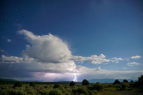 A dramatic night sky with clouds and lightning illuminating the landscape, surrounded by mountains and sagebrush.