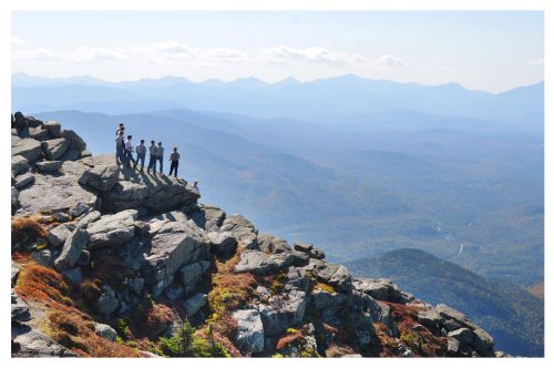 A group of hikers stands on a rocky ledge, overlooking a vast mountain landscape with rolling hills and blue skies.