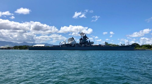 A large battleship docked in a harbor, surrounded by water and under a clear blue sky with scattered clouds.