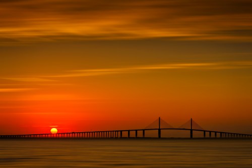 A sunset casts vibrant orange hues over a bridge silhouetted against the horizon.