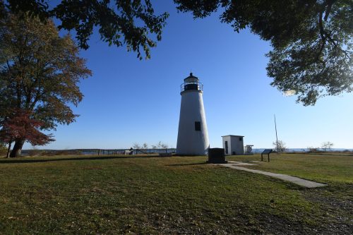 A white lighthouse stands on a grassy area with trees, under a clear blue sky, overlooking a body of water.