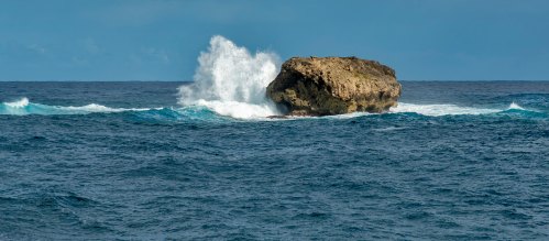 A rocky outcrop in the ocean with waves crashing against it under a cloudy sky.