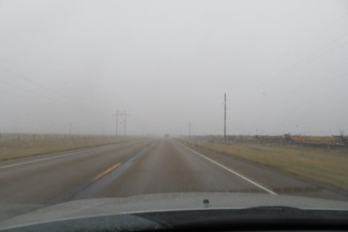 A foggy road stretches ahead, with low visibility and power lines lining the sides. Rain is visible on the windshield.