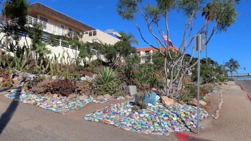Colorful painted rocks decorate a landscaped area with cacti and trees, beside a road and residential buildings.