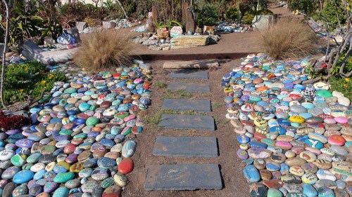A colorful stone pathway lined with painted rocks leads to a garden area with shrubs and decorative stones.