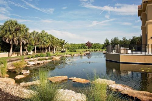 A serene landscape featuring a pond surrounded by rocks, palm trees, and a sculpture under a clear blue sky.