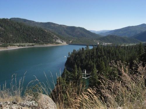 A serene landscape featuring a blue lake surrounded by green forests and mountains under a clear blue sky.