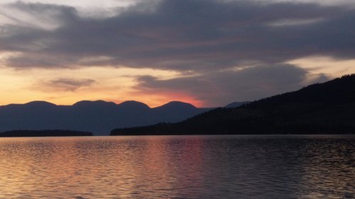A serene lake at sunset, with mountains silhouetted against a colorful sky and calm water reflecting the hues.