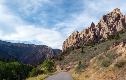 A winding road alongside rocky cliffs and green vegetation under a blue sky with scattered clouds.