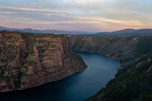 A serene river winding through steep, rocky cliffs under a colorful sky at dusk. Lush greenery surrounds the water.