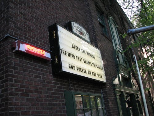 A movie theater marquee displays titles: "After the Wedding," "The Wind That Shakes the Barley," and "Buy Volver on DVD Here."