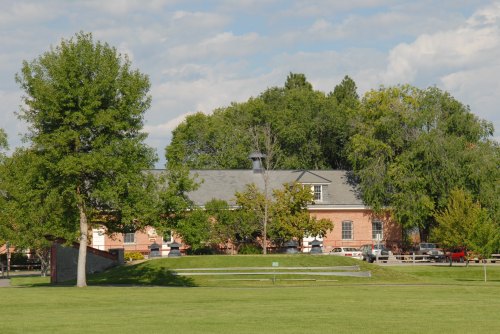 A brick building surrounded by trees and a grassy area, with cars parked nearby under a partly cloudy sky.