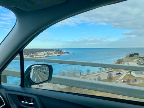 View from a car window overlooking a calm lake, with a shoreline and industrial buildings in the distance under a cloudy sky.