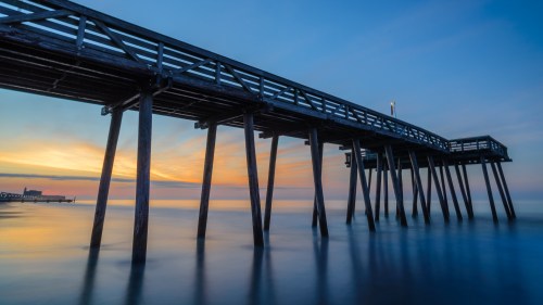 A wooden pier extends over calm waters at sunset, with soft colors in the sky reflecting on the surface.