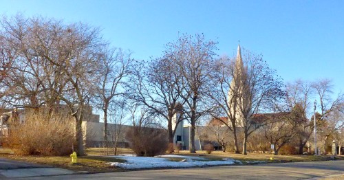 A winter scene featuring bare trees, a church spire, and a clear blue sky. Snow patches are visible on the ground.