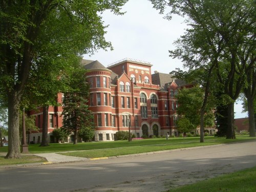 Historic red brick building surrounded by trees and green grass, with a clear sky in the background.