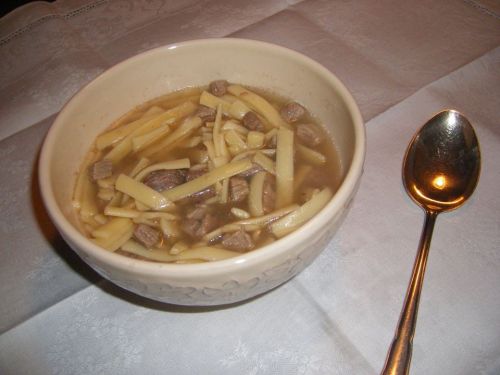 A bowl of soup with noodles and pieces of meat, accompanied by a silver spoon on a white tablecloth.
