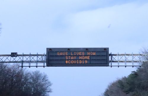 Digital highway sign displaying: "SAVE LIVES NOW STAY HOME #COVID19" against a cloudy sky.
