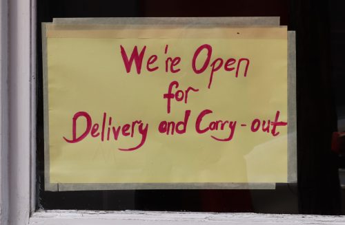 A bright yellow sign in a window reads, "We're Open for Delivery and Carry-out" in bold pink letters.
