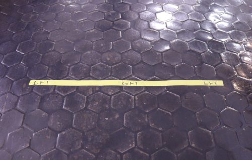 Yellow tape marked "6FT" on a hexagonal tiled floor, indicating a distance guideline.