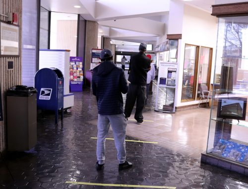 Two people stand in a lobby, maintaining distance, with a postal service box and informational displays in the background.