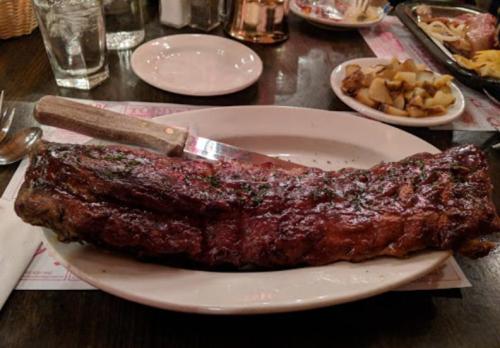 A large, glazed rack of ribs on a white plate, accompanied by a side of diced potatoes.
