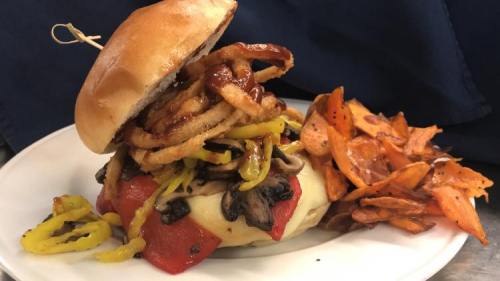 A gourmet burger topped with sautéed mushrooms, peppers, and crispy onions, served with sweet potato fries.