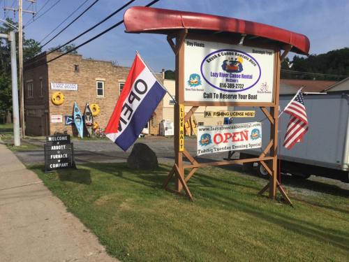 Sign for Lazy River Canoe Rental with flags, indicating open status, and nearby buildings and kayaks in the background.