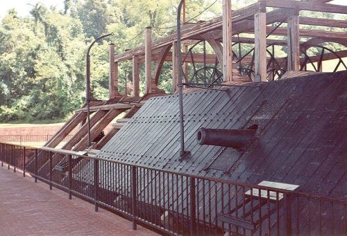 Historic wooden structure with a sloped black roof, featuring large wheels and a cannon, surrounded by greenery.