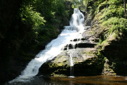 A cascading waterfall flows over rocky steps, surrounded by lush green foliage and a serene pool at the base.