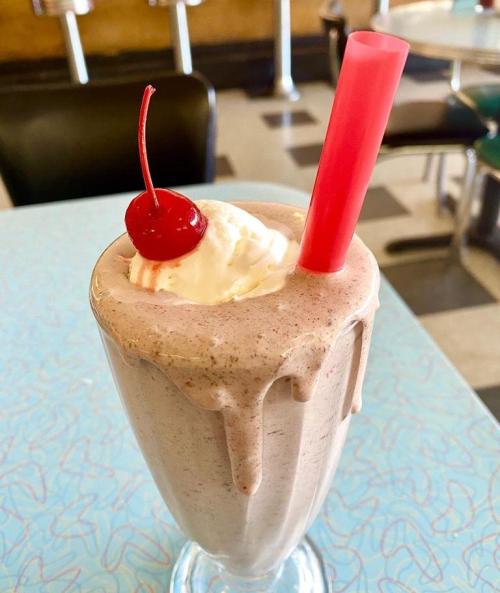 A thick milkshake topped with whipped cream and a cherry, served in a tall glass with a pink straw.