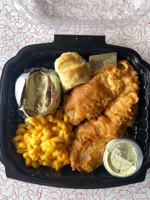 A takeout container with fried fish, macaroni and cheese, a baked potato, a biscuit, and a side of sauce.